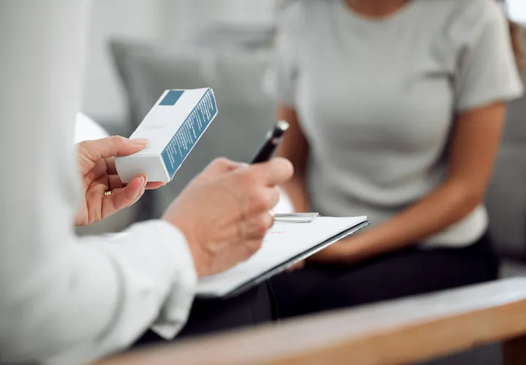 A doctor holds a box of medication in their hand while writing on their clipboard. A patient sits across from them.