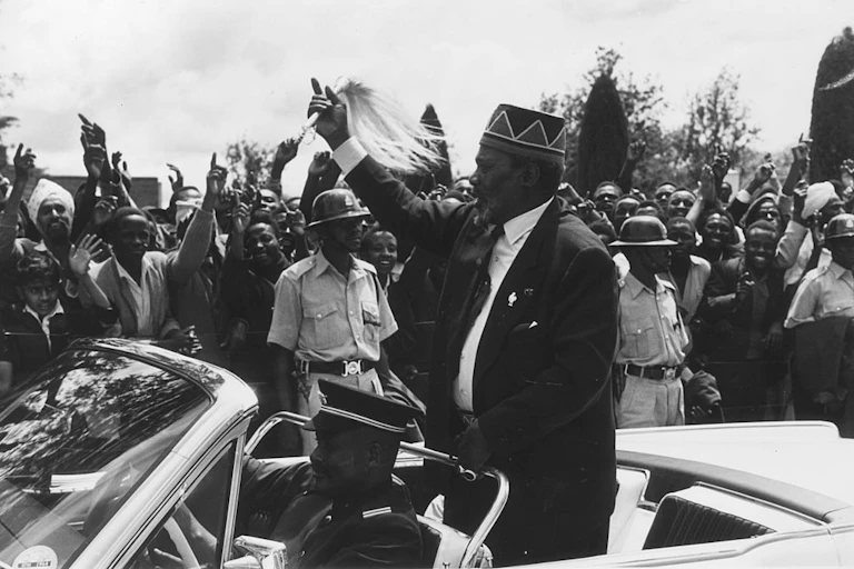 A black and white image of a man waving a flywhisk while holding up one finger and gesturing at a crowd. He is standing at the back of a vehicle driven by a man in military uniform. A cheering crowd of men and women waves back while holding up their index fingers