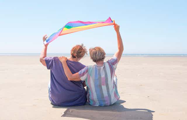 View from behind of an older lesbian couple sitting on a beach with their arms around each other, holding a pride flag above their heads