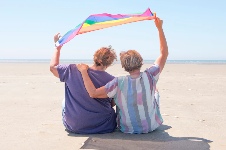 View from behind of an older lesbian couple sitting on a beach with their arms around each other, holding a pride flag above their heads