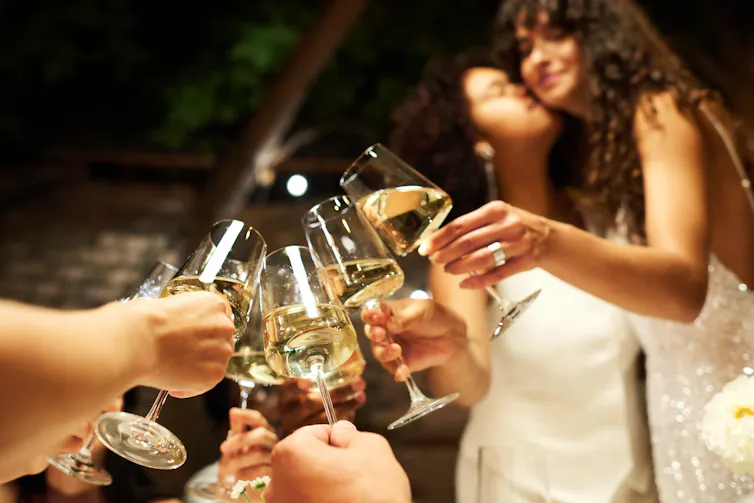 Photo of champagne flutes raised in a toast, while two brides embrace in the background