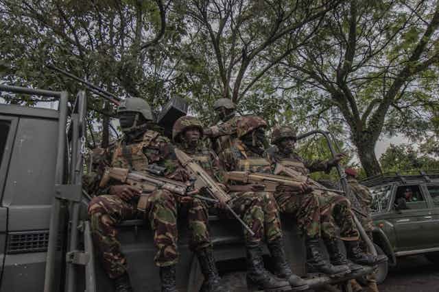 Men wearing military uniform and holding guns sit in the back of an open pick-up truck