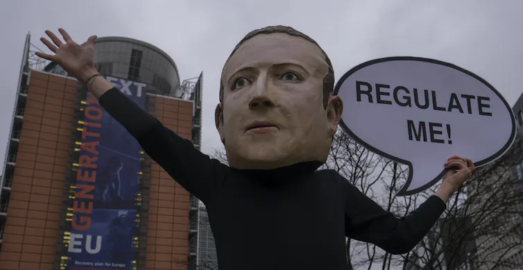 A person wearing an oversized papier mâché Mark Zukerberg head holding up a sign that reads 'regulate me'.