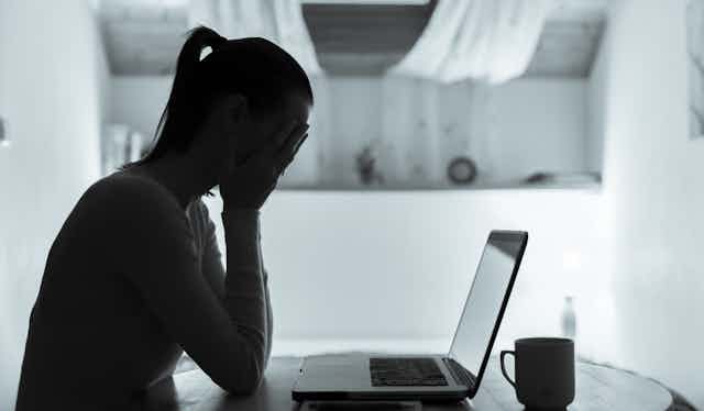A woman seen in silhouette sitting at a table in front of an open laptop with her head in her hands.