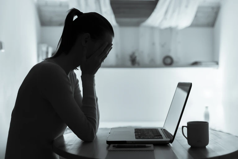 A woman seen in silhouette sitting at a table in front of an open laptop with her head in her hands.