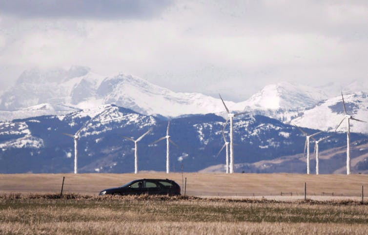 A black car drives past a wind farm. Snow capped mountains are in the background.