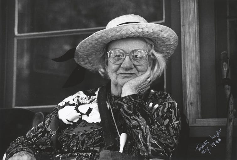 An elderly woman in a broad-brimmed hat and glasses, chin in hand
