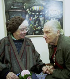 Two older women holding hands in front of a painting: one talking, one intently listening