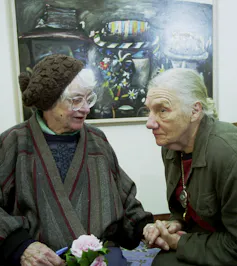 Two older women holding hands in front of a painting: one talking, one intently listening