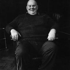 Les Murray: a large man dressed in black, sitting in a chair, smiling
