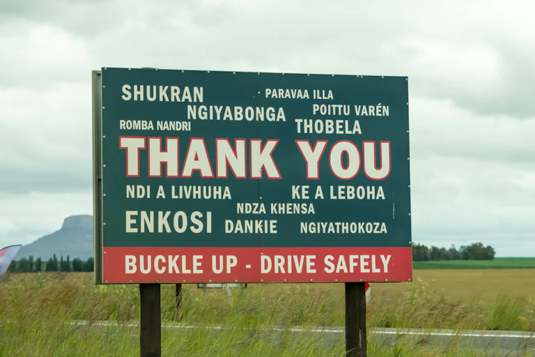 A roadside sign implores passersby to "buckle up, drive safely" in a variety of languages