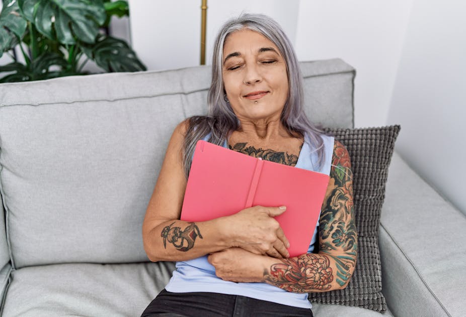 Woman with grey hair sitting on a sofa and hugging a red bound book.