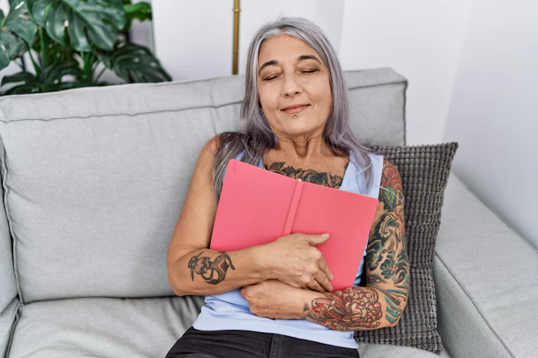 Woman with grey hair sitting on a sofa and hugging a red bound book.