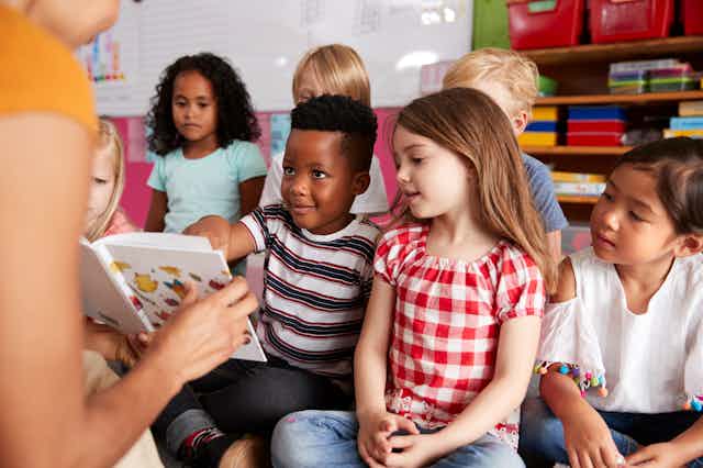 Un niño señala algo en un libro ante la mirada de la profesora que sujeta el libro y otros compañeros de clase.