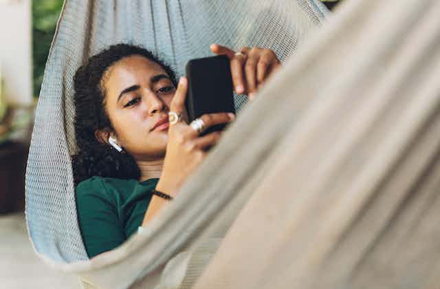 A young woman laying in a hammock, looking at her mobile phone while wearing earbuds