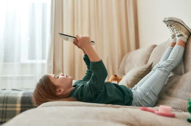Una niña observa una tableta tumbada en el sofá y con las piernas apoyadas en el respaldo.
