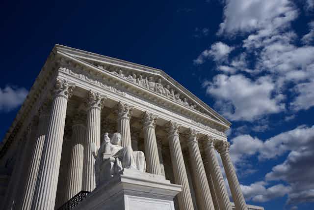 A view of the Supreme Court from the outside, with tall white pillars, against a blue sky. 