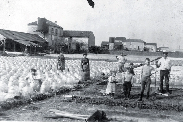 Photo en noir et blanc, montrant une dizaines de personnes (hommes, femmes et enfants), debout en train de poser au milieu des cloches et des cultures, avec les batiments de l'exploitation en arrière-plan