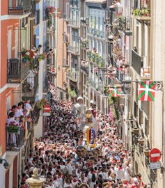 Una calle estrecha abarrotada de gente de fiesta mientras más gente observa desde los balcones de los edificios.