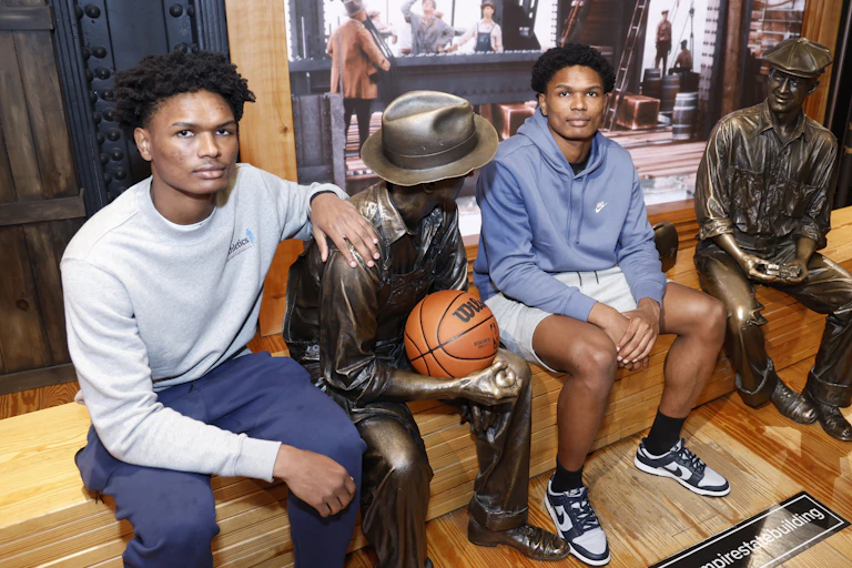 Two young men sit on a bench next to a statue with a basketball in the statue's hands.
