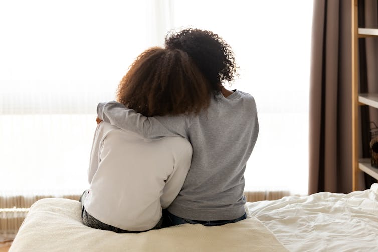 A woman with her arm around a teen, sitting on the edge of a bed, seen from behind