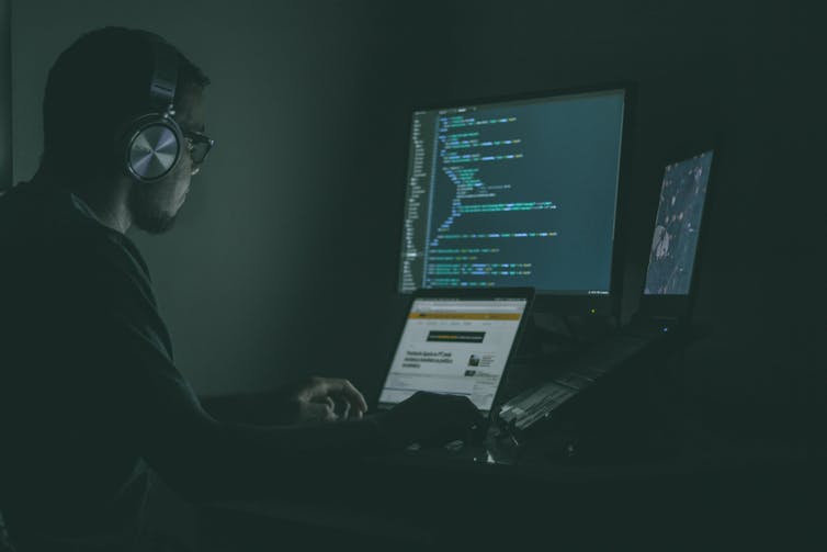 a man sits in the dark in front of a laptop and additional screen. he is wearing headphones