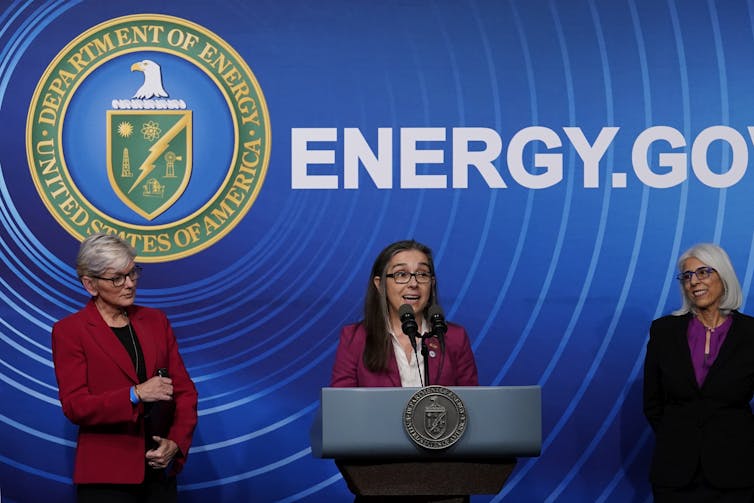Three women people in business attire stand in front of a screen that says 'Energy.Gov.' The women in the centre speaks into a microphone from behind a podium.