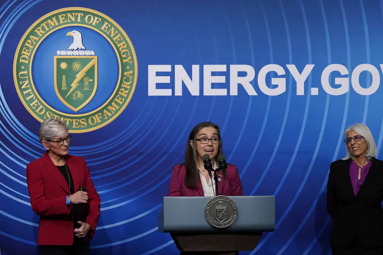 Three women people in business attire stand in front of a screen that says 'Energy.Gov.' The women in the centre speaks into a microphone from behind a podium.