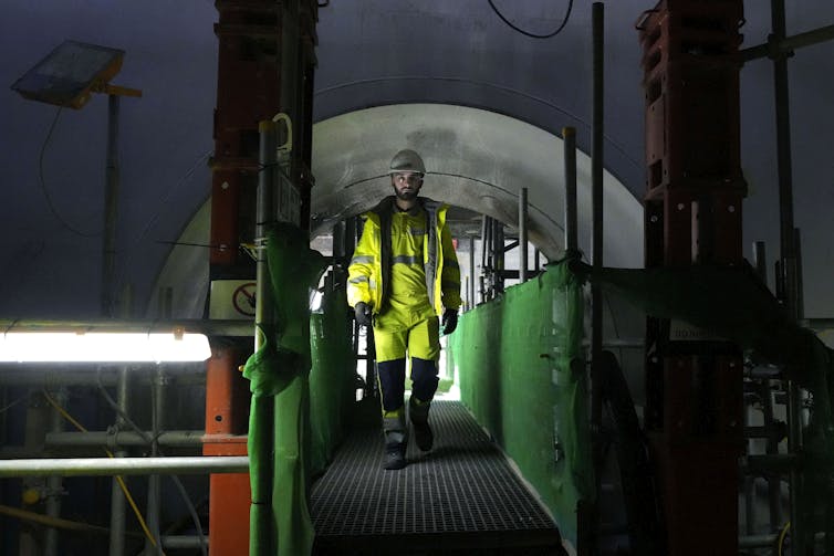 A person in a bright yellow construction uniform and hardhat walks on a small bridge inside a nuclear reactor that is under constriction.