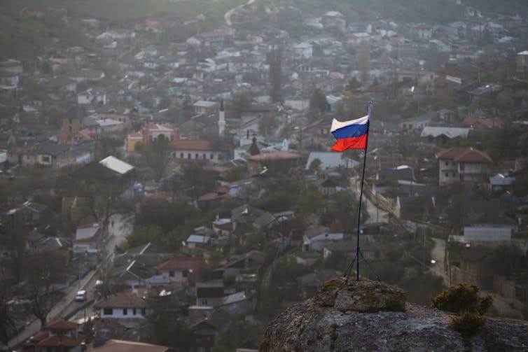 A red white and blue flag flies on a hilltop overlooking a city.