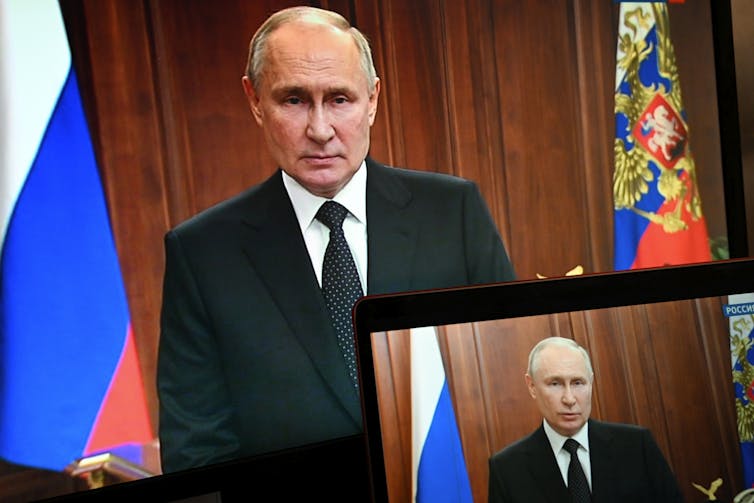 A balding man is seen on two screens with flags behind him.