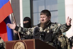 Un hombre barbudo en uniforme de combate extiende los brazos mientras habla detrás de un podio.