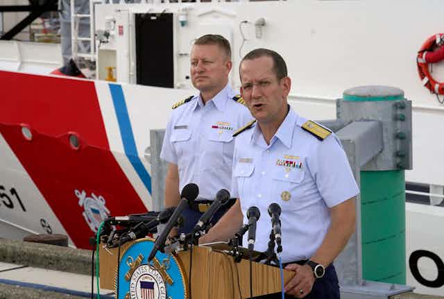 Two men in light blue polo ships with ornamental shoulder pieces address people off camera from behind a podium.