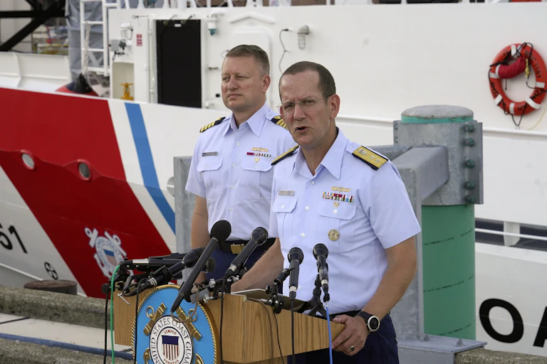 Two men in light blue polo ships with ornamental shoulder pieces address people off camera from behind a podium.