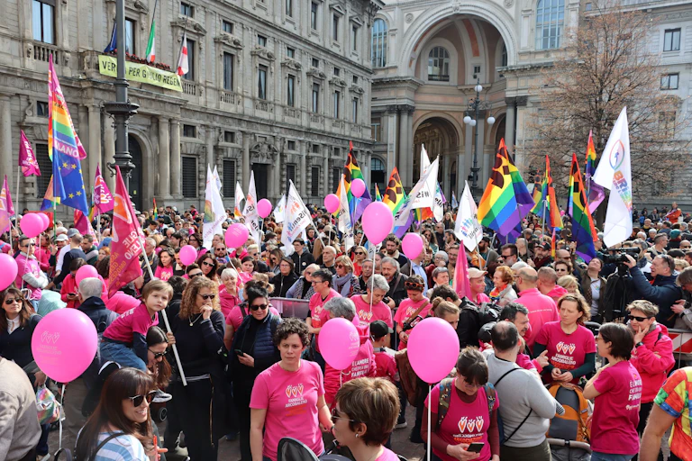 A group of people carrying LGTBQ flags protest in an old square.