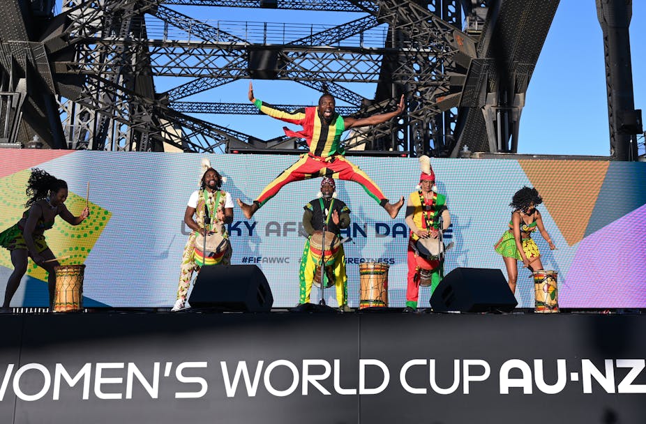 Dancers jumping and playing instruments on a stage that says the words 'women's World Cup Au-NZ'.