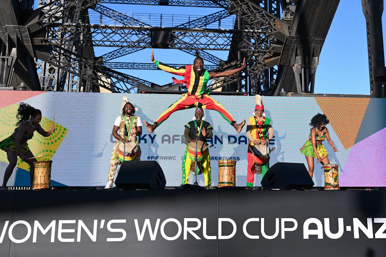 Dancers jumping and playing instruments on a stage that says the words 'women's World Cup Au-NZ'.
