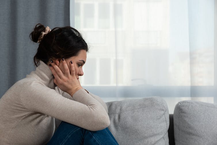 A woman sitting on a couch, holding her head between her hands.