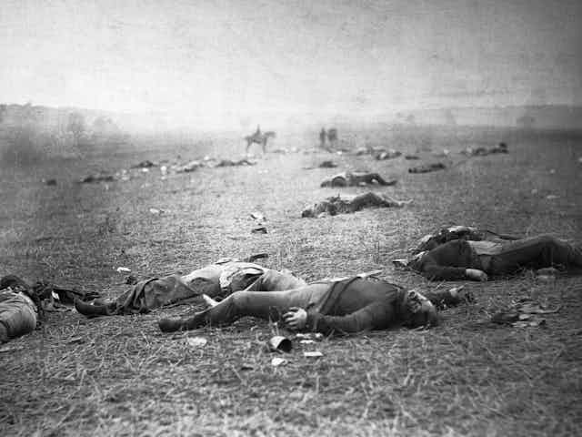 A black and white photo depicts a body-strewn field, with a person on horseback visible in the distance.