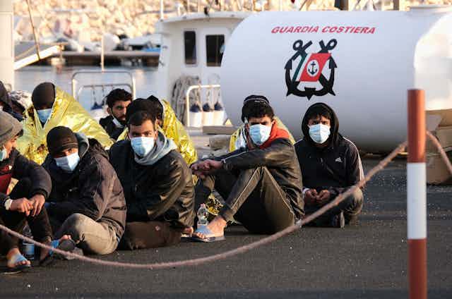 Migrants sitting on the ground wearing masks look at the camera.