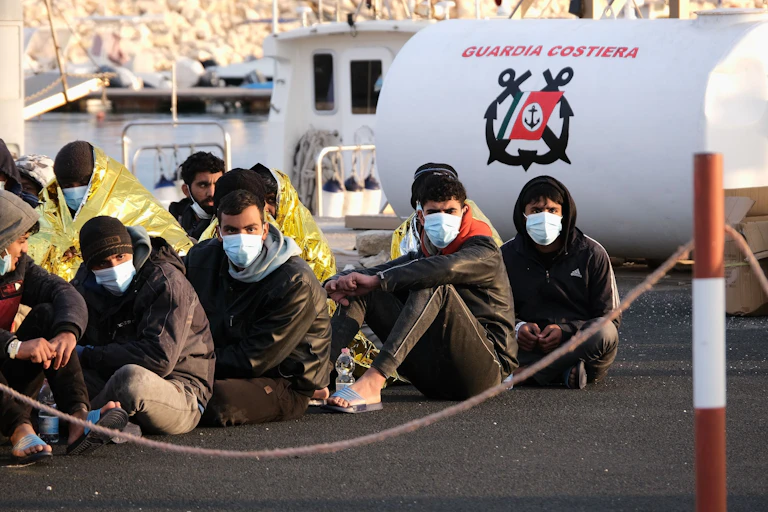 Migrants sitting on the ground wearing masks look at the camera.