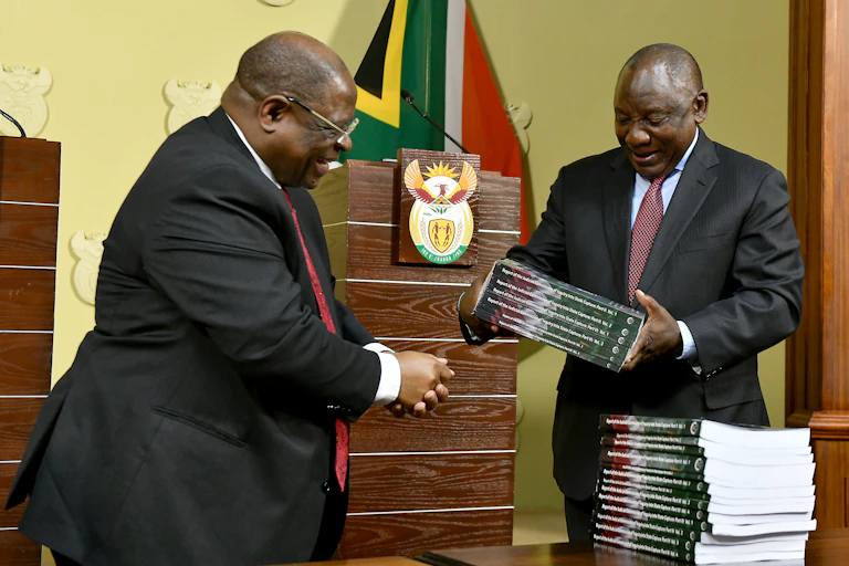 A smiling man wearing a suit and tie receives a voluminous report from another on his right.