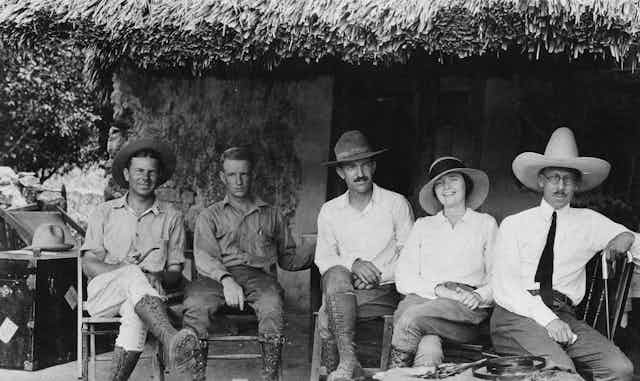 Cuatro hombres y una mujer vestidos de exploradores están sentados en el exterior de una cabaña mientras miran a cámara.