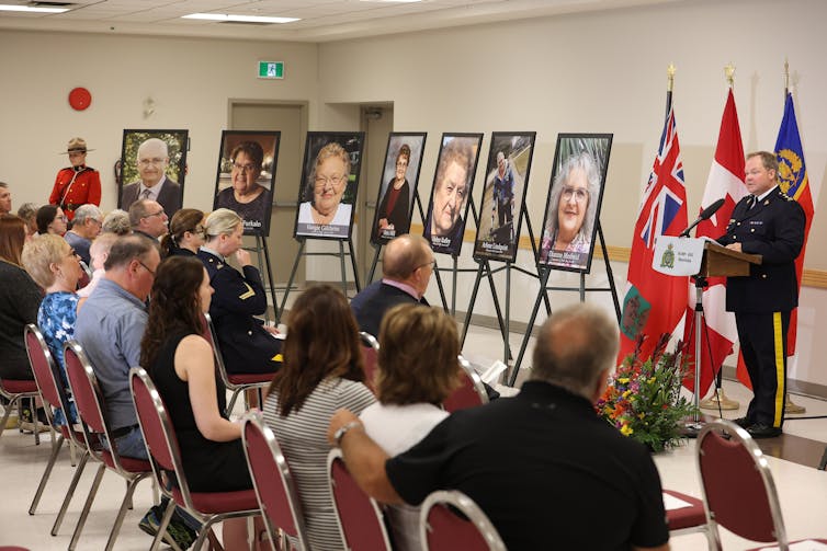 People sit in a room with large portraits. An RCMP officer stands at a podium addressing them.