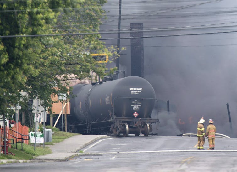 Firefighters stand looking at clouds of black smoke and the back of a rail car in the middle of the road.