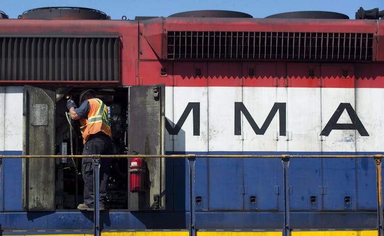 A man in an orange hazard vest looks at the engine of a red white and blue train that says MMA on its side.