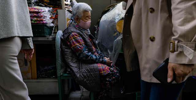 An old woman in a mask sits in a chair while people walk past her.