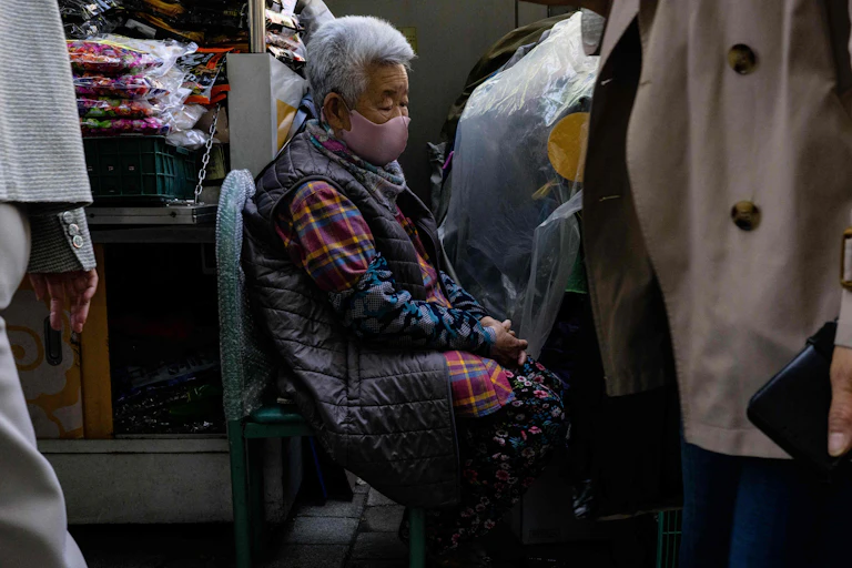 An old woman in a mask sits in a chair while people walk past her.