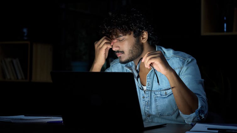 A stressed man in front of a laptop places his fingers on his forehead