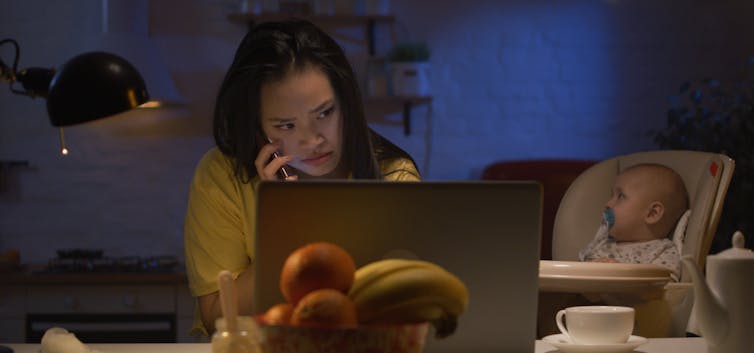A woman working on a laptop with a baby beside her in a high chair